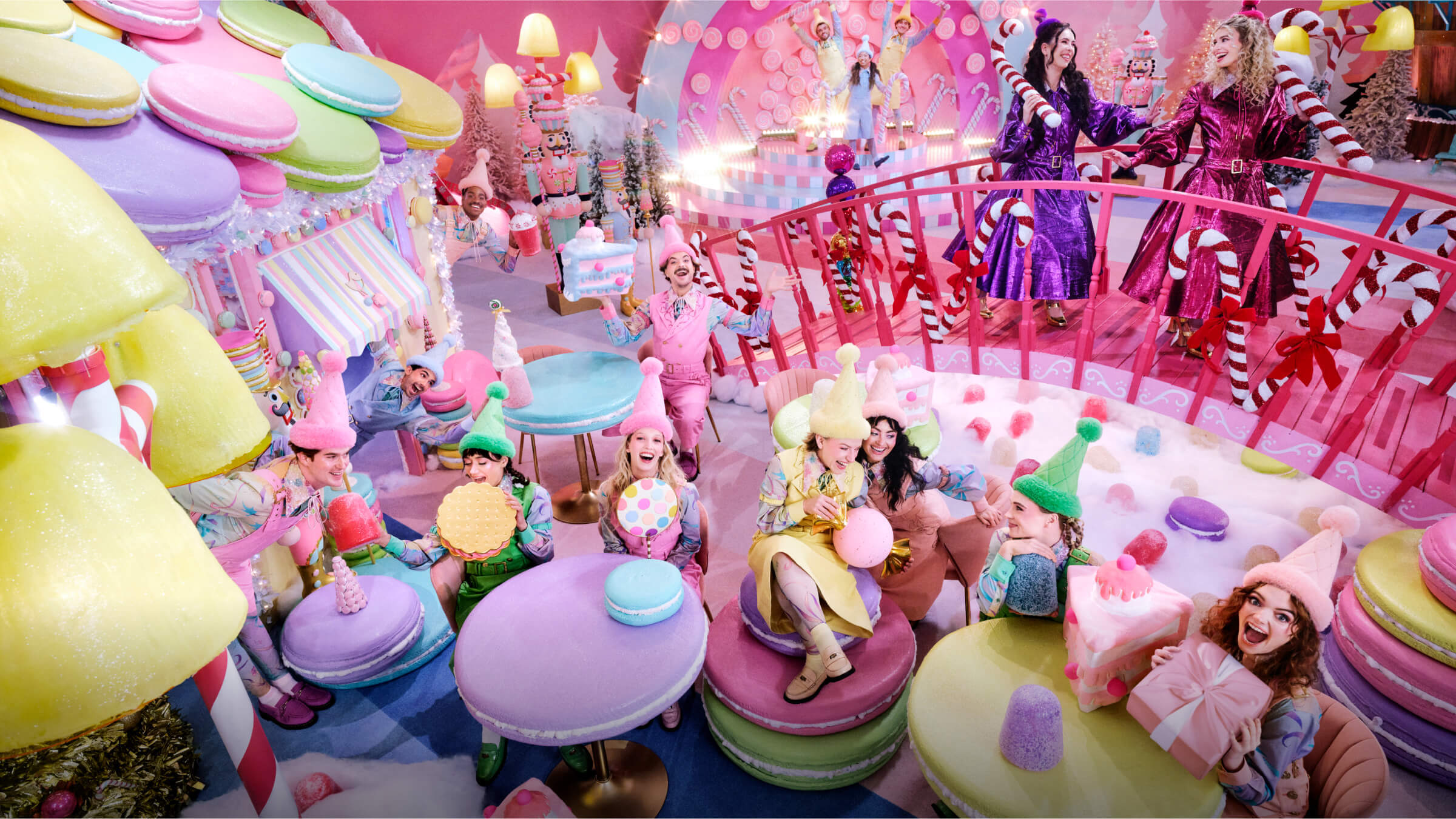 A colorful candy-themed scene with people dressed as elves and characters, surrounded by giant sweets like macarons and candy canes, creating a playful, whimsical atmosphere